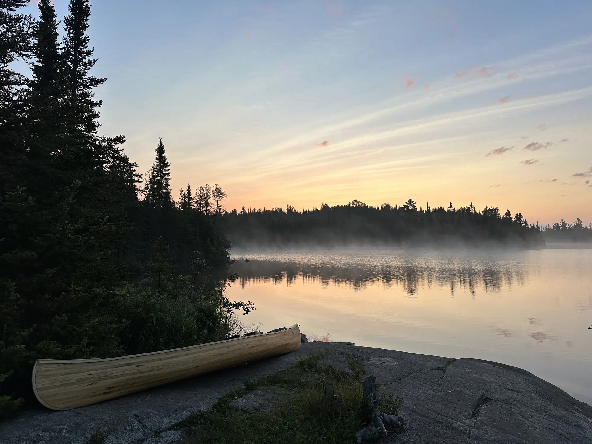 A wood-strip canoe pulled up on a rock ledge beside a misty BWCA lake at sunrise